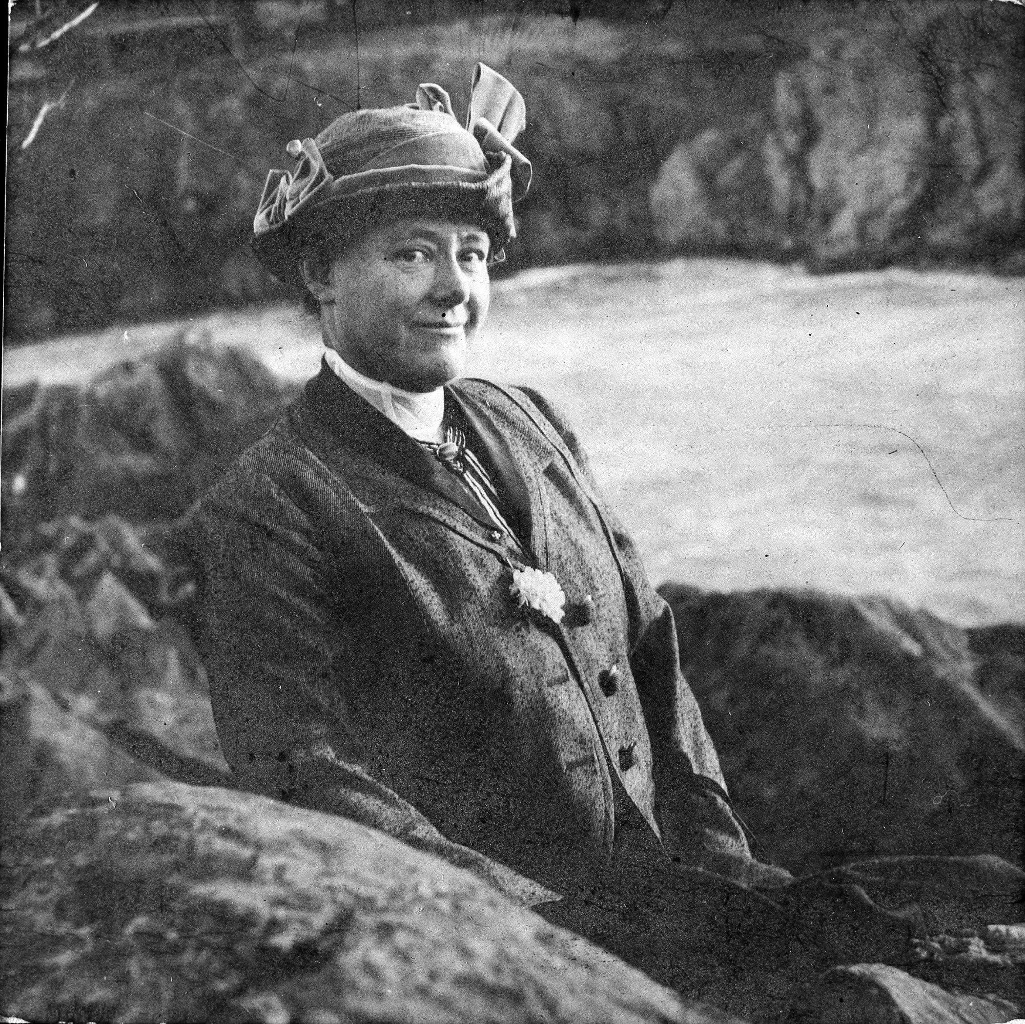 Black and white vintage portrait of a woman wearing a dark tailored jacket, high collar, and decorative hat, posed outdoors against a rocky natural background.