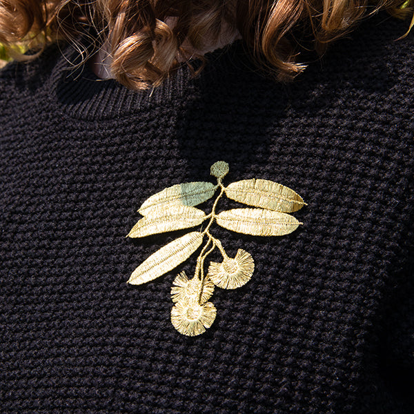A close-up of a person wearing a black textured knit sweater with a metallic gold embroidered leaf and seed branch brooch pinned to the chest.