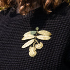 A close-up of a person wearing a black textured knit sweater with a metallic gold embroidered leaf and seed branch brooch pinned to the chest.