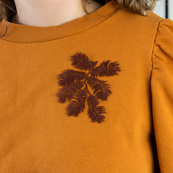 Close-up of a woman wearing a mustard yellow sweatshirt with a dark brown embroidered leaf design on the chest.
