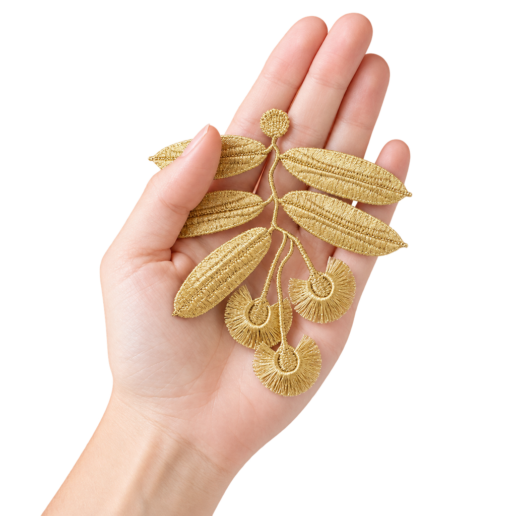 A hand holding a gold embroidered leaf and seed branch applique against a plain white background.