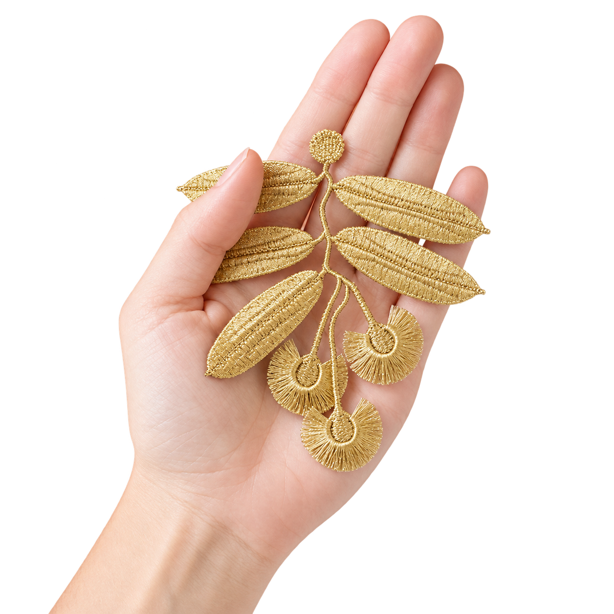 A hand holding a gold embroidered leaf and seed branch applique against a plain white background.