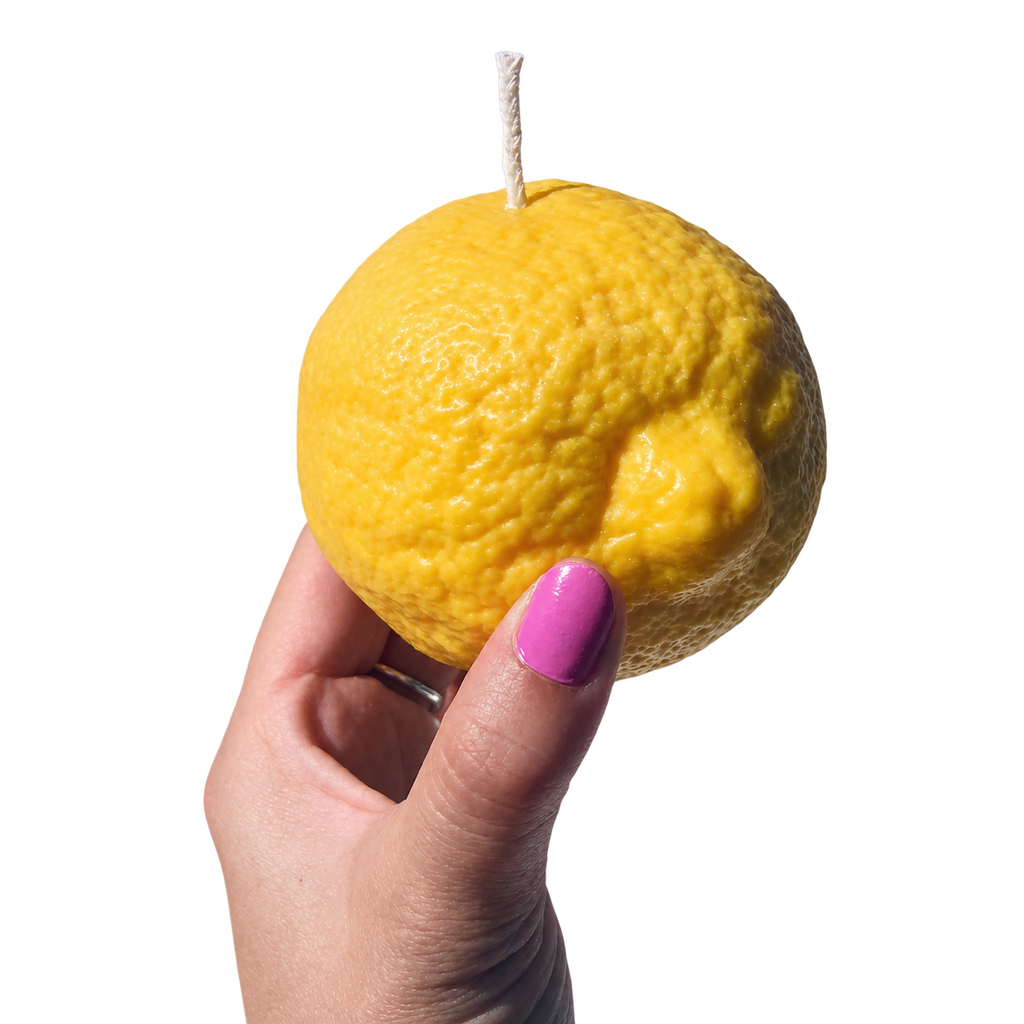 A hand with pink nail polish holds a novelty candle shaped like a yellow lemon, featuring a visible wick on top, against a plain white background.