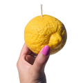 A hand with pink nail polish holds a novelty candle shaped like a yellow lemon, featuring a visible wick on top, against a plain white background.