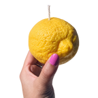 A hand with pink nail polish holds a novelty candle shaped like a yellow lemon, featuring a visible wick on top, against a plain white background.