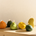 Five fruit-shaped wax candles with lit flames are arranged on a light wood surface against a plain beige wall. The candles include an orange, dark green, light green pear, a yellow lemon, and a yellow mango.