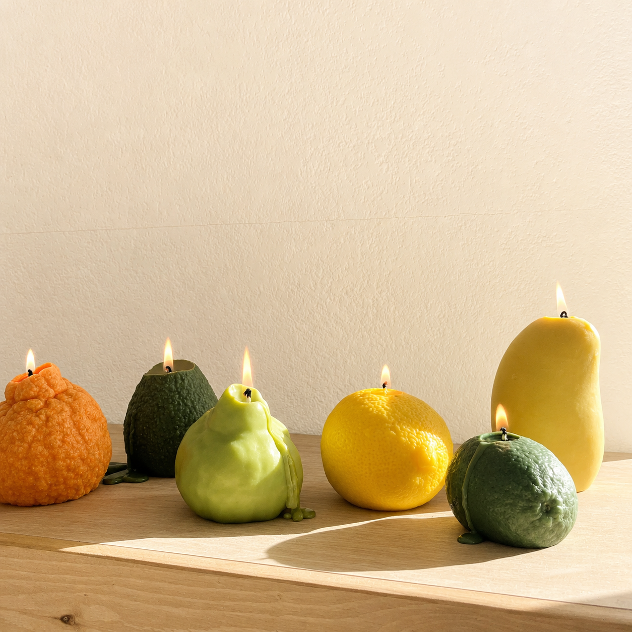 Five fruit-shaped wax candles with lit flames are arranged on a light wood surface against a plain beige wall. The candles include an orange, dark green, light green pear, a yellow lemon, and a yellow mango.