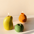 Three sculptural lit candles shaped like a yellow mango, an orange citrus peel, and a green lime, arranged on a white surface against a warm beige background.