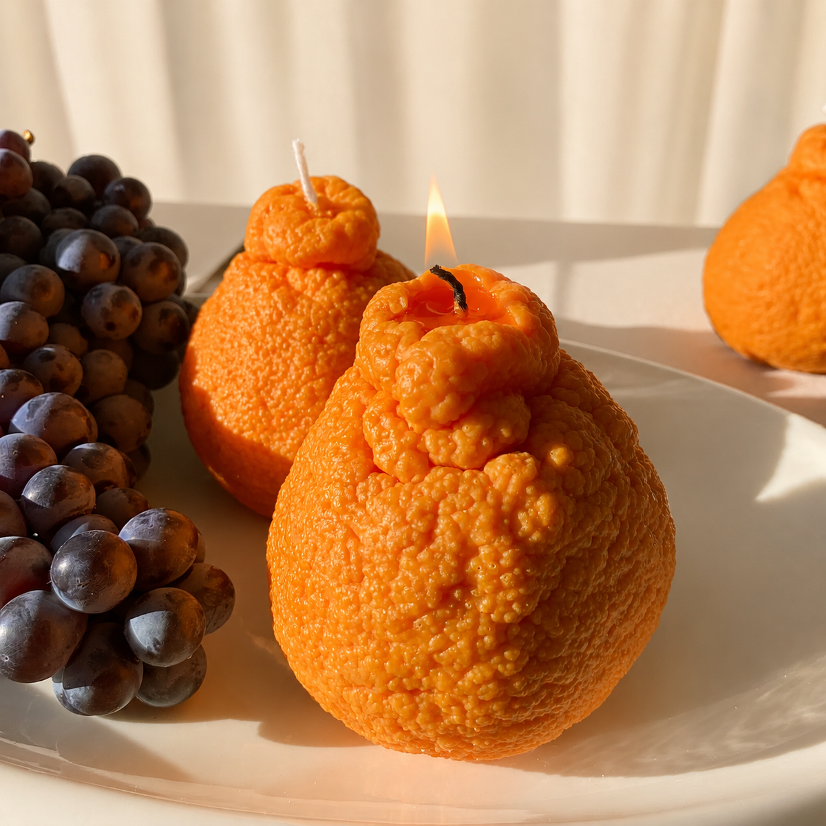 Three sculpted orange candles shaped like oranges, displayed on a white plate with a lit candle in the foreground, alongside a bunch of dark grapes and soft sunlight filtering through curtains.