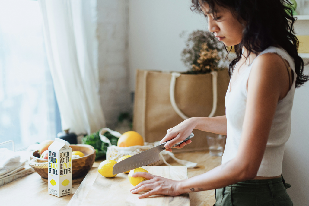 Woman slicing lemons on a wooden cutting board with a carton of milk and oranges in the background.