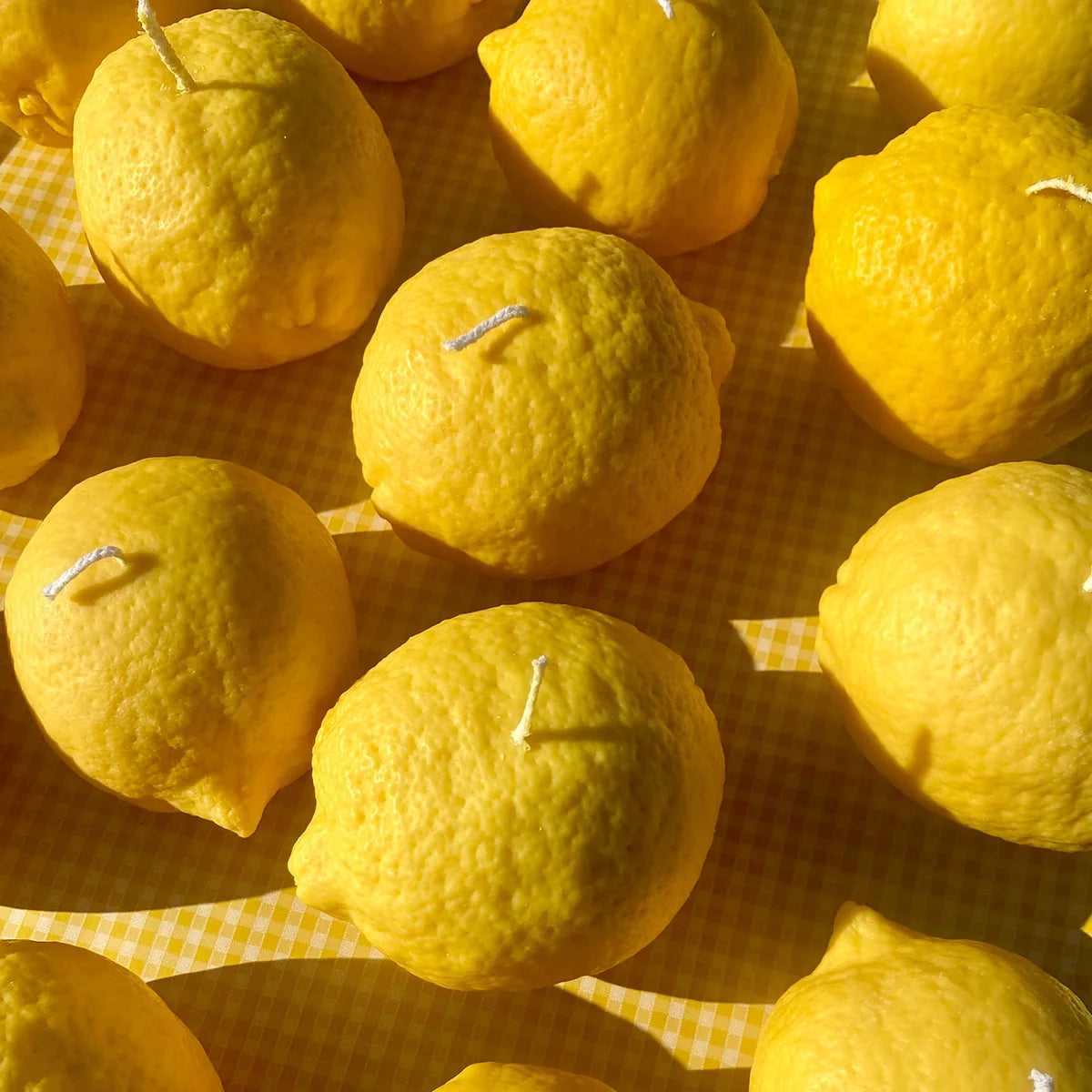 A close-up arrangement of bright yellow lemons on a yellow-and-white checkered surface, with small white wicks visible on several lemons, suggesting they are candles.