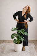 Woman in a black dress standing next to a potted plant in a room with a light-colored wall.