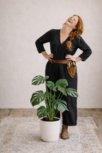 Woman in a black dress standing next to a potted plant in a room with a light-colored wall.