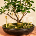 A miniature garden tree display in a dark oval planter, featuring green leaves, mossy ground, and small gold decorative accessories including a bench, bicycle, fence, lamp post, and hanging ornaments.