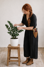 Woman in a black dress tending to a potted ZZ plant on a wooden stool against a plain wall.